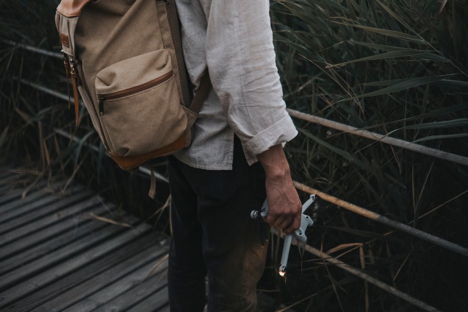 A man wearing a backpack stands on a wooden walkway holding a drone, surrounded by tall gr