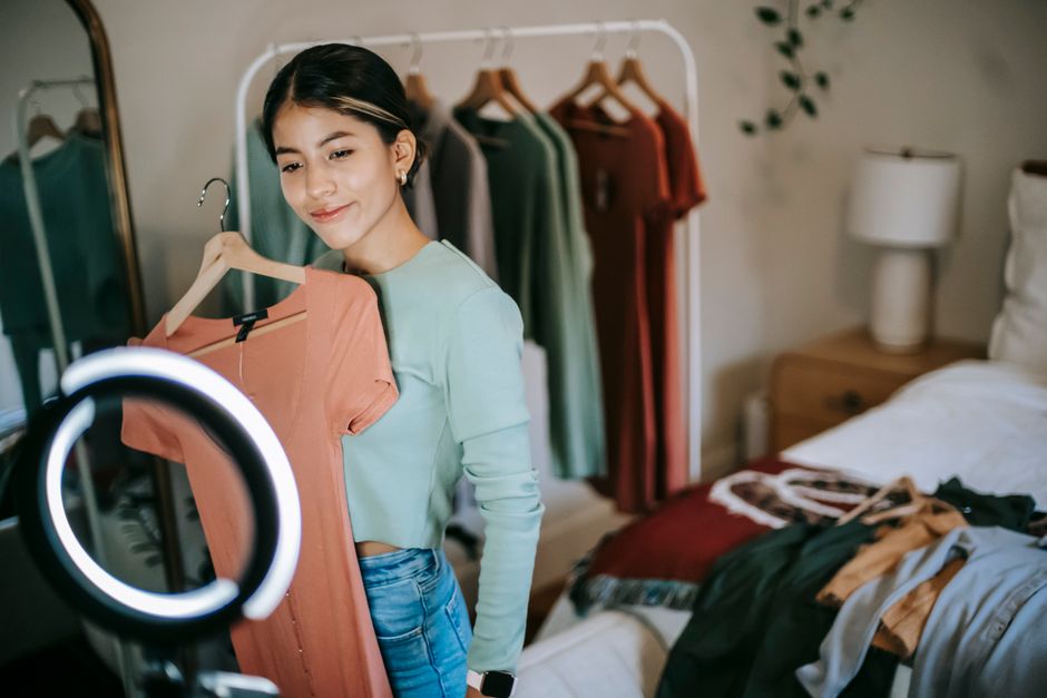 Happy young ethnic female blogger standing in bedroom and demonstrating pink dress on hanger while filming video on smartphone with ring lamp near bed and mirror near wardrobe