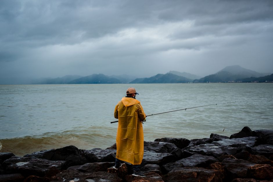 Back view of unrecognizable male fisher in yellow raincoat standing on rocky coast near wa