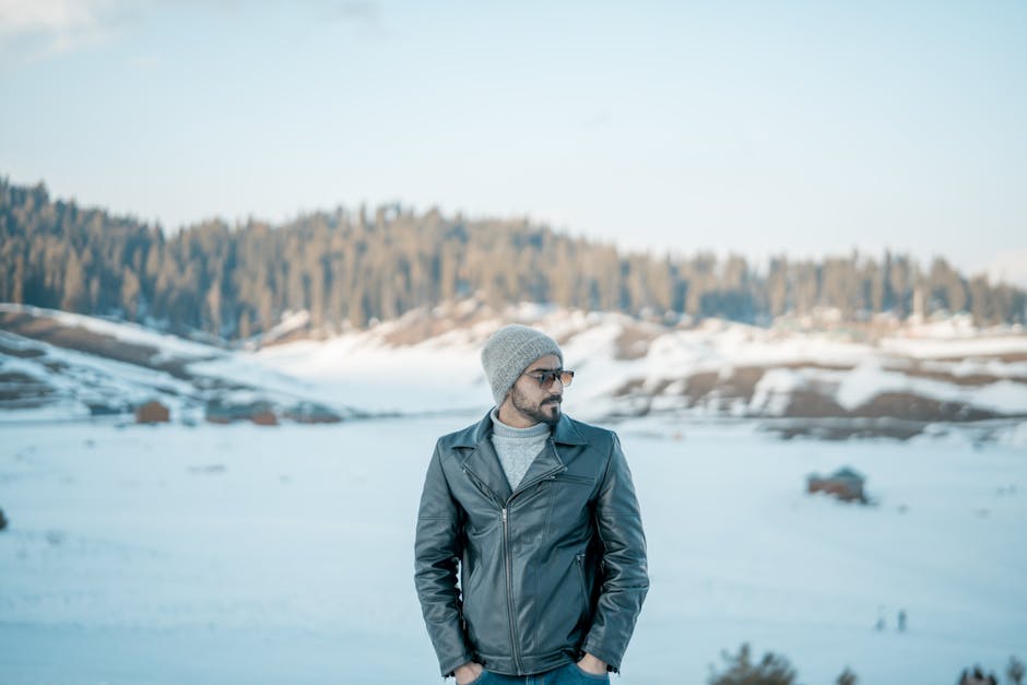 Man in a leather jacket at Gulmarg, Kashmir, surrounded by snow-covered mountains under a clear sky.