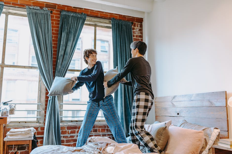 Two teenage boys enjoying a playful pillow fight in a cozy bedroom setting.