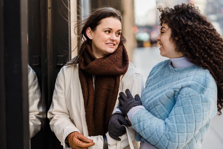 Delighted young diverse female friends in trendy warm clothes smiling and looking at each other while chilling on city street on cold day
