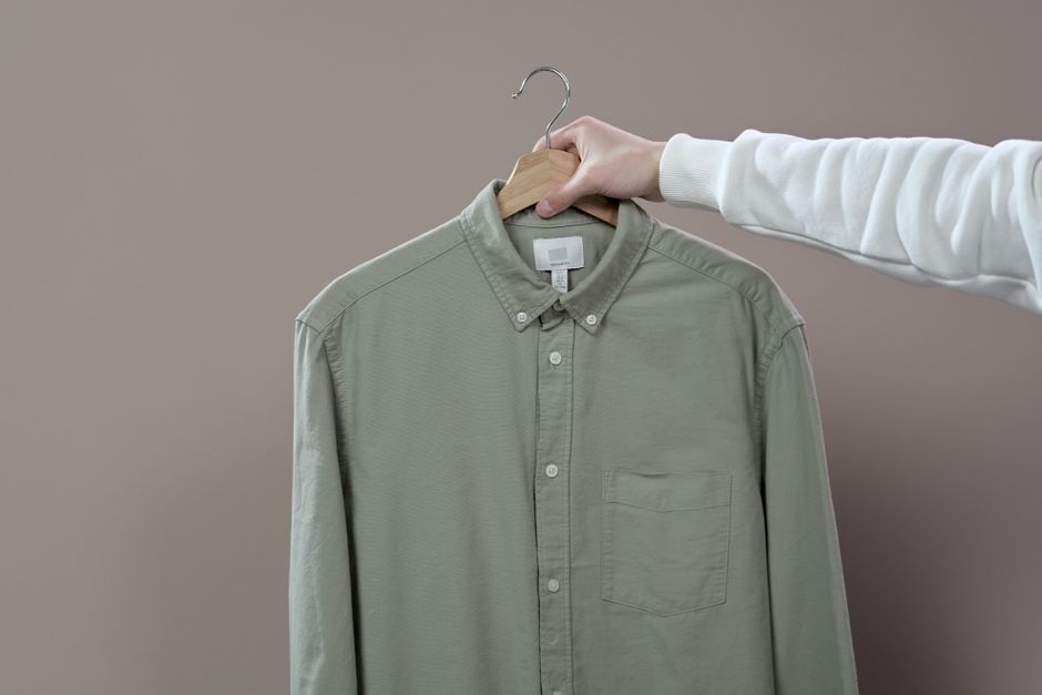 A hand holding a green button-up shirt on a hanger in a minimalist studio setting.