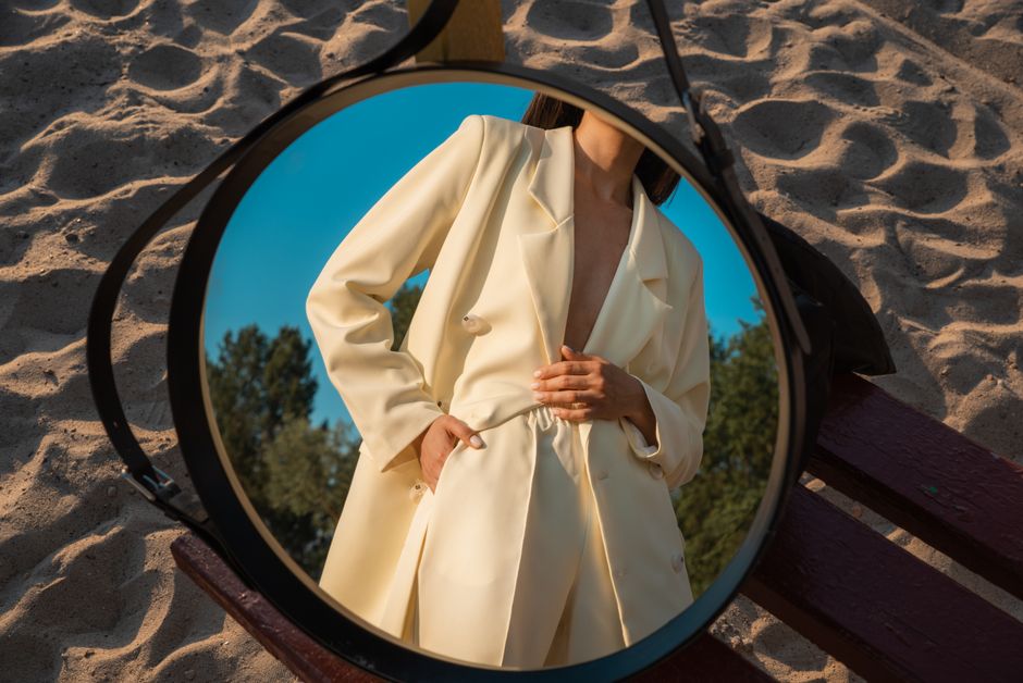Fashionable woman in white suit reflected in a mirror on a sandy beach setting.