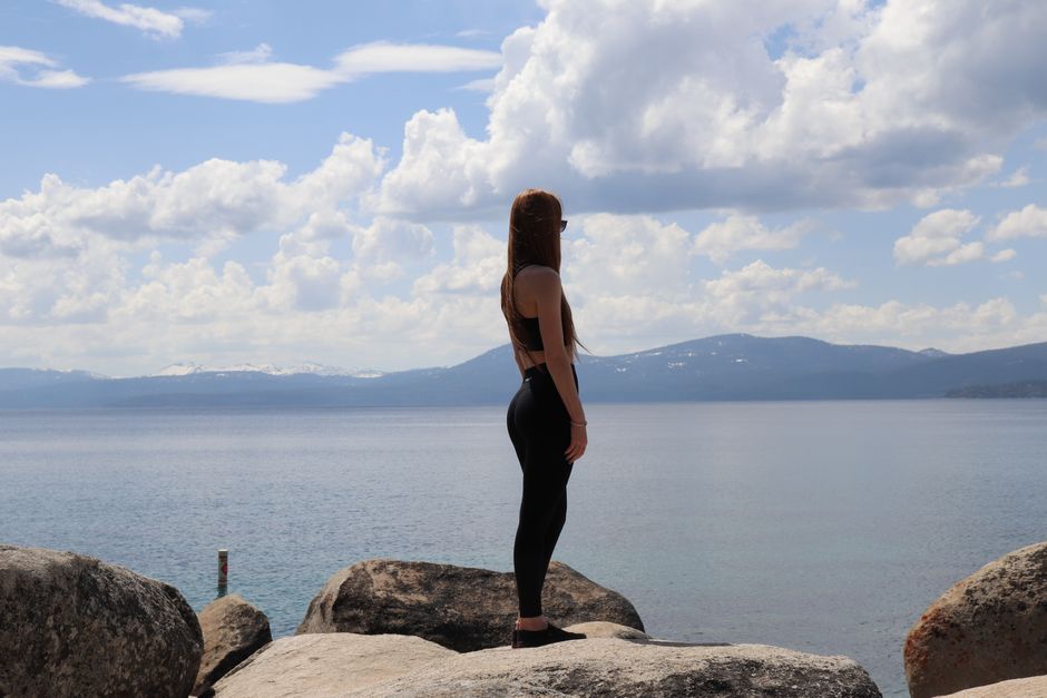 A woman in sportswear stands on rocks by a calm lake under a partly cloudy sky.