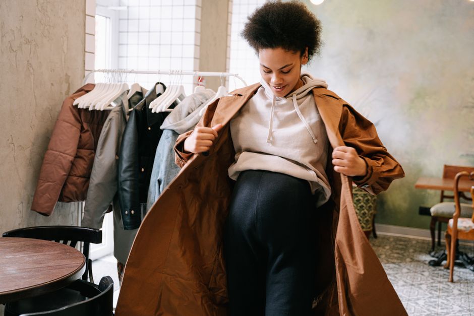 Pregnant woman in stylish coat adjusting outfit indoors near clothes rack.