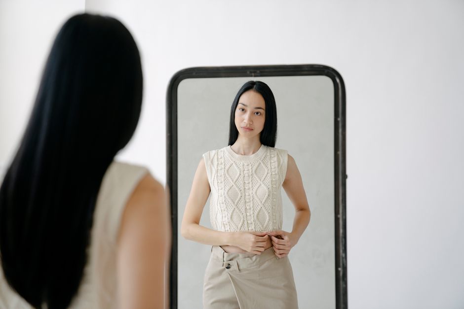Back view of young Asian female in knitted sleeveless shirt reflecting in mirror in light studio