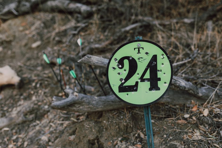 A circular archery target sign with bullet holes, surrounded by arrows outdoors.