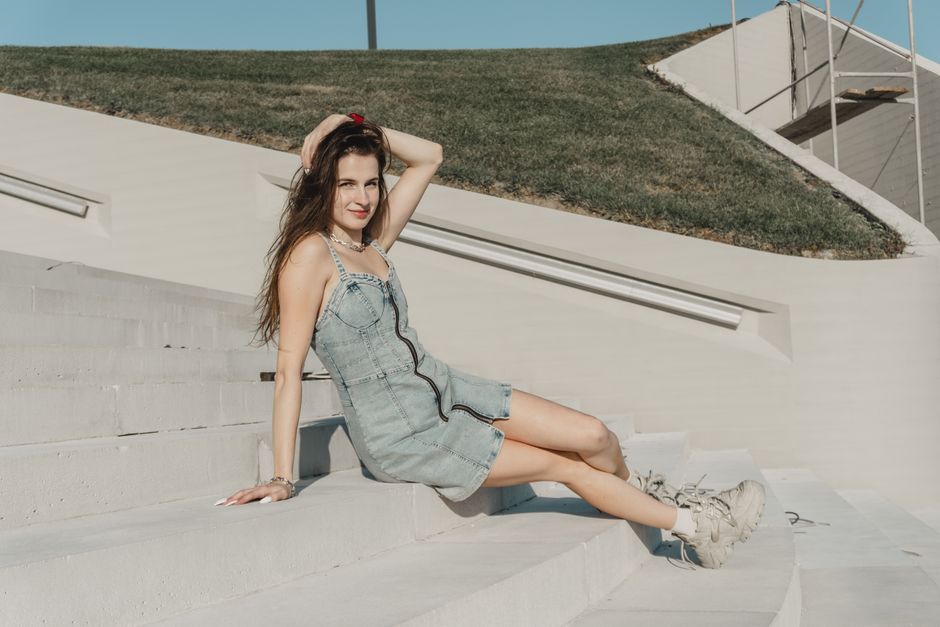 Stylish young woman in a denim dress poses on outdoor concrete steps under clear skies.