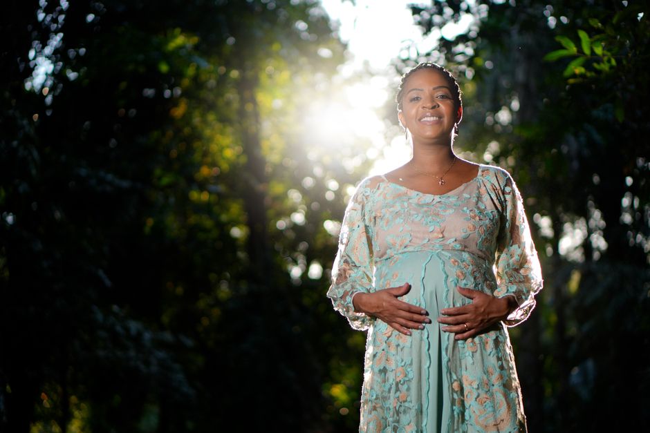 Smiling pregnant woman in a forest with sunlight filtering through trees.