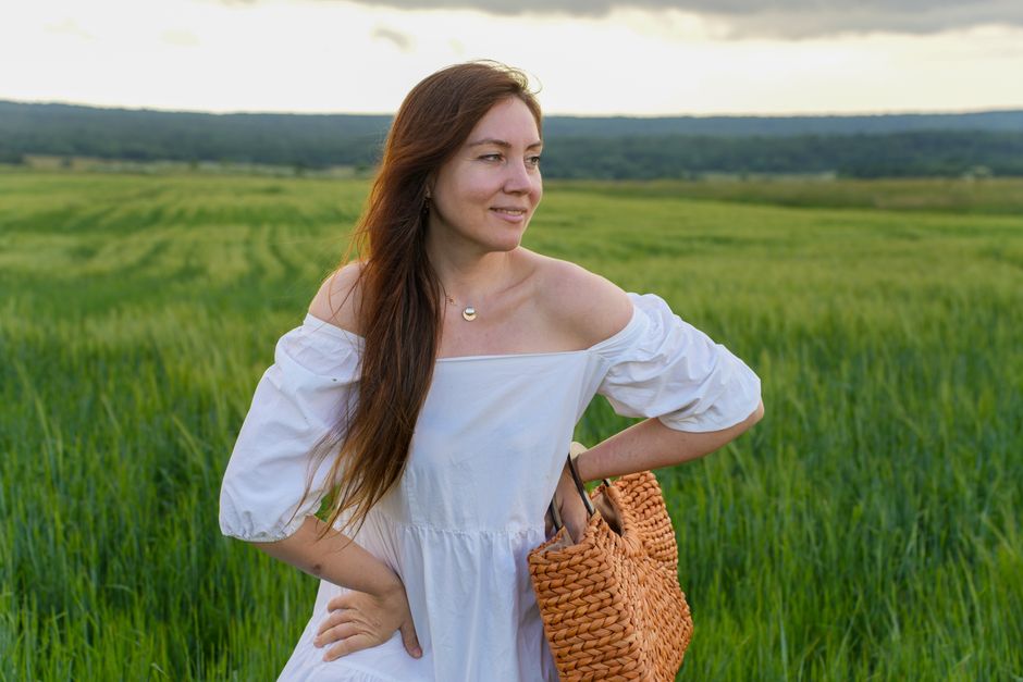 A woman in a white dress stands in a vibrant green field, holding a woven basket, under a cloudy sky.