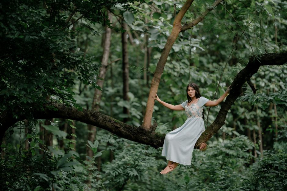 A woman in a white dress sitting on a tree in a lush green forest.