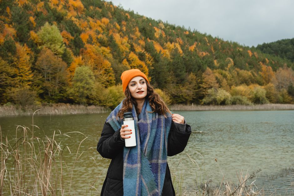 Young woman in autumn attire with thermos by scenic lake, vibrant fall foliage.